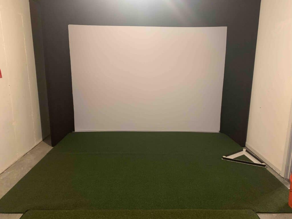 Golf simulator, white backdrop with green turf in front.