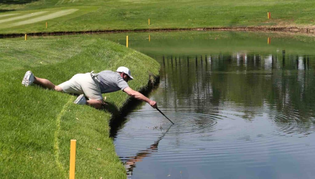 A golfer, after hitting his ball in the water, tries to retrieve it using a golf club.